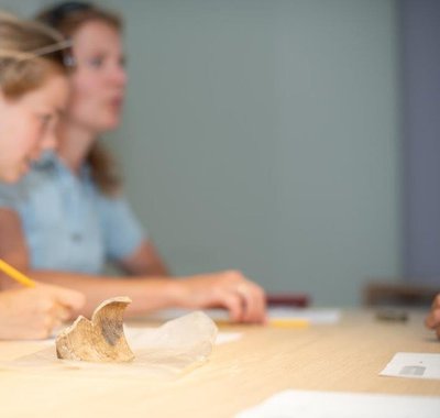 Twee meisjes zitten aan tafel en luisteren aandachtig naar uitleg over een archeologische vondst dat bij hen op tafel ligt.