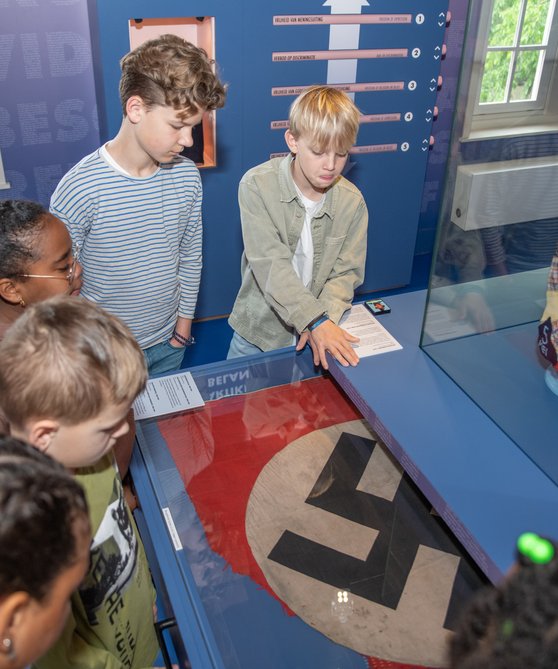 Een groepje kinderen staat voorovergebogen rond een vitrine met daarin een hakenkruisvlag in Hof van Nederland.