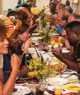 Aan een lange Keti Koti Tafel is een gevarieerd gezelschap druk met elkaar in gesprek. Er staat eten en drinken op tafel.